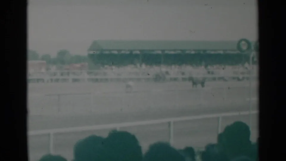 Home-movie frame of a Cheyenne horse race, seen past foreground spectators toward the track rail and green-roofed grandstand.