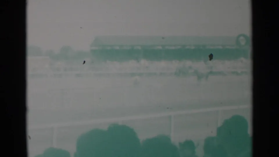 Soft 1960 8mm view of spectators at a Cheyenne racetrack, with a long grandstand and track rail visible beyond foreground heads.