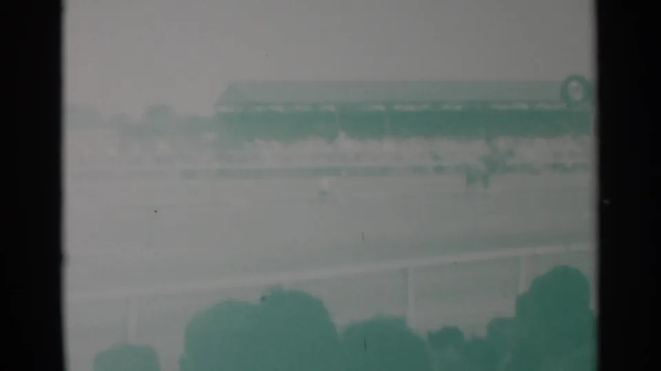 Cheyenne horse race frame with distant horses near the rail, foreground spectator silhouettes, and a crowded grandstand under hazy light.