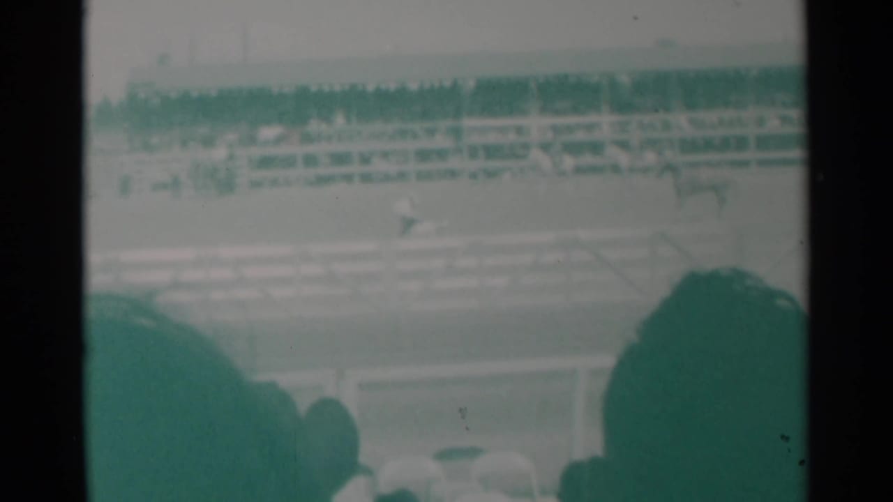 Arena railings, distant riders and crowded bleachers seen over foreground spectators.