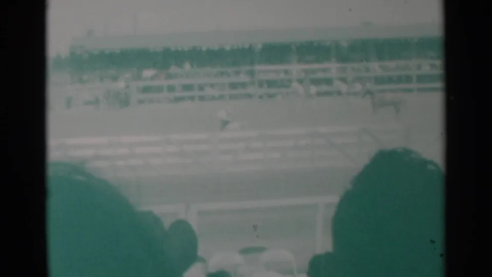 Cheyenne rodeo footage shows distant horseback activity across the arena, with bleachers and dark spectator silhouettes in front.