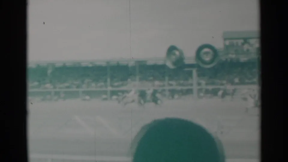 Wide 1960 Cheyenne rodeo arena view with distant riders, grandstands, and foreground crowd obstruction.