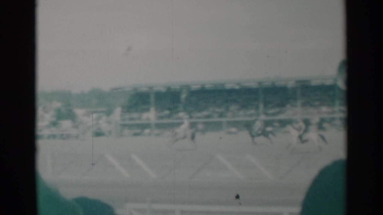1960 Cheyenne rodeo view with bronco riders crossing the arena before crowded grandstands in soft green-tinted 8mm footage.