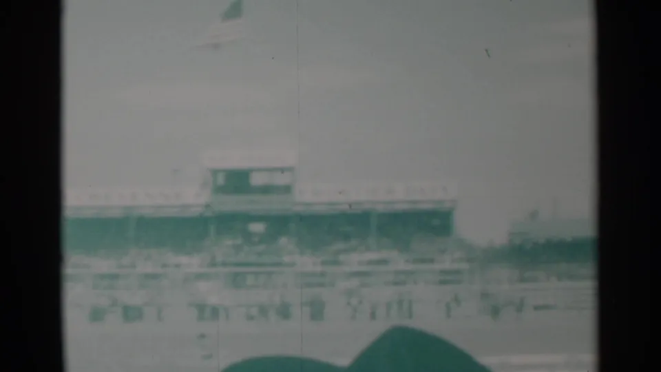 Faded 8mm frame showing the Cheyenne rodeo grandstand, arena fencing, and a crowd gathered near the rail.