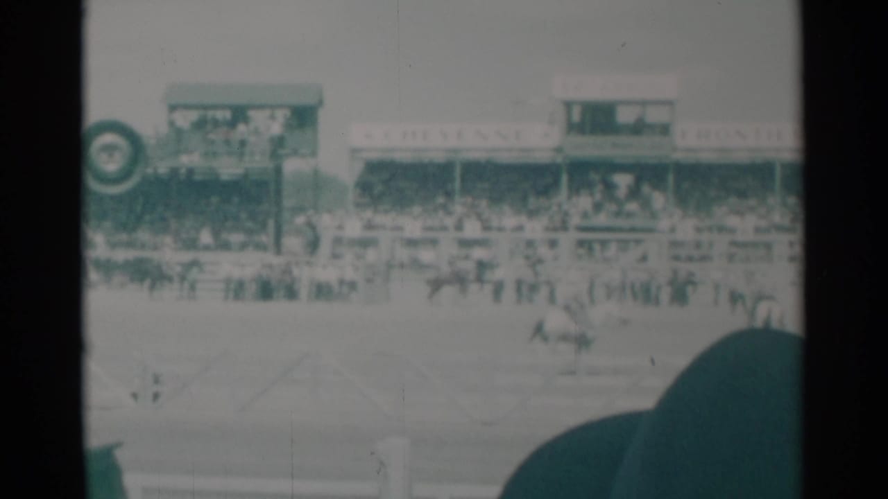 Rodeo arena with riders, grandstand spectators, signage, and a dark hat in the foreground.
