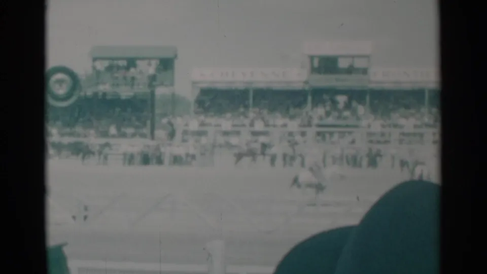 1960 Cheyenne rodeo view from the stands, with riders in the arena, spectators under Frontier Days signage, and a dark hat partly blocking the frame.