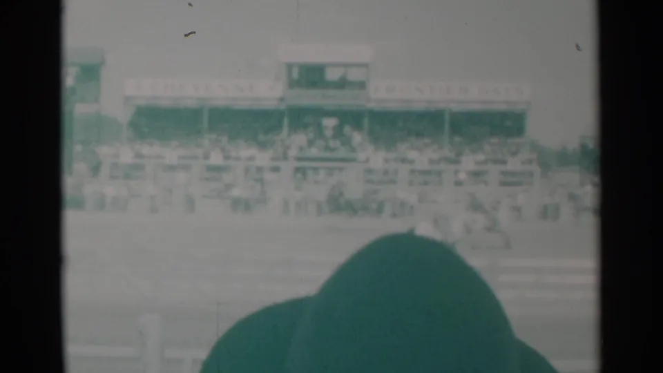 Home-movie view of the Cheyenne rodeo arena, with the grandstand signage and spectators visible beyond a large foreground hat.