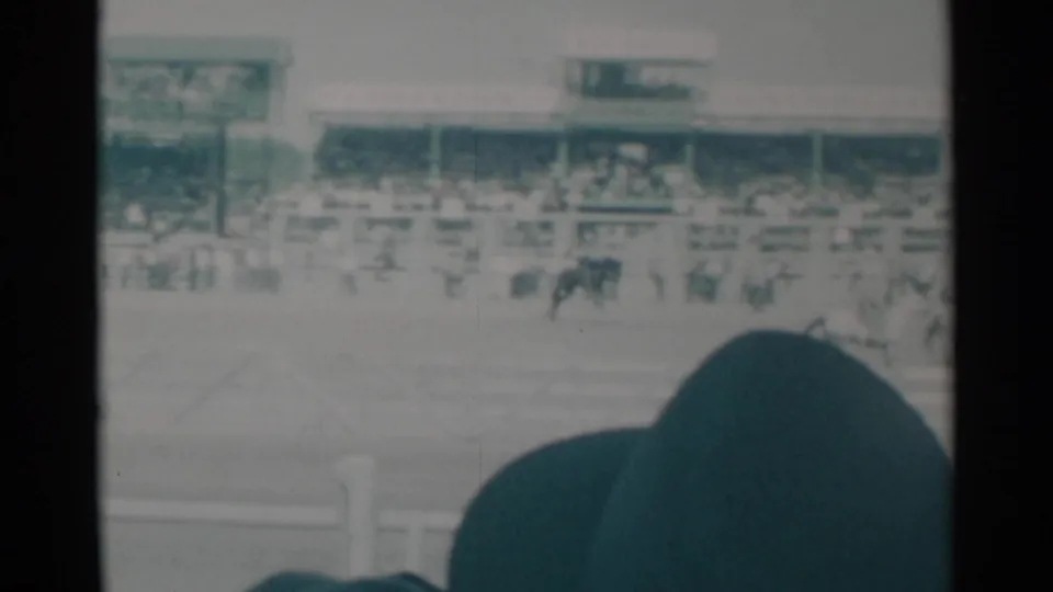 1960 Cheyenne, Wyoming frame with a rider moving across the arena, grandstand crowds, signage, and a foreground spectator hat.