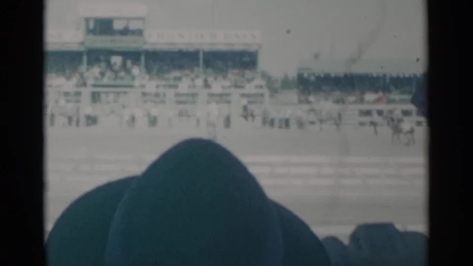 Home-movie view of the Cheyenne rodeo grandstand and track, partly obscured by a nearby spectator in the lower frame.