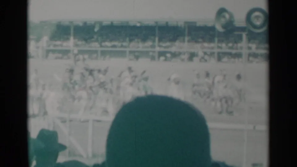 A spectator-shot 8mm view of horses, riders, and a grandstand crowd in Cheyenne, partly blocked by a foreground audience member.