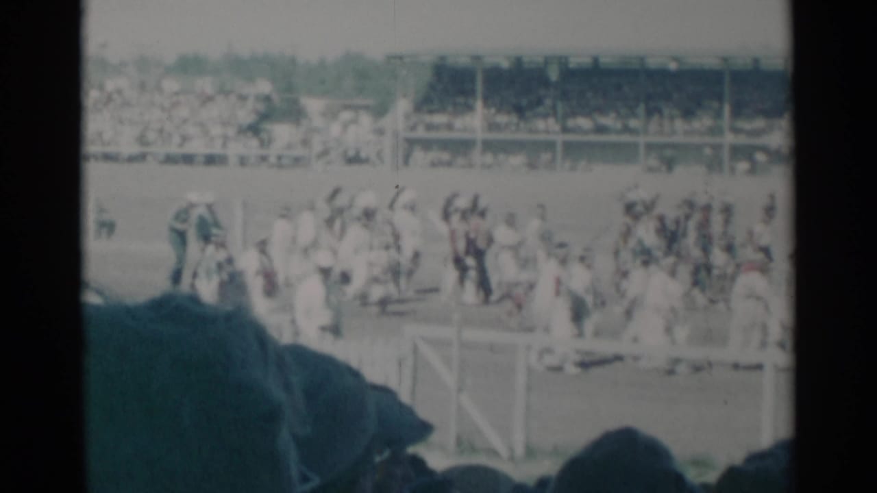Crowd crossing a field near grandstands, seen past dark spectator silhouettes.