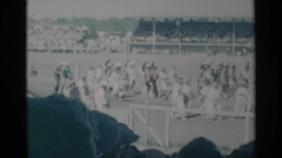 1960 Cheyenne home-movie frame showing people moving across an event field with filled grandstands behind them.