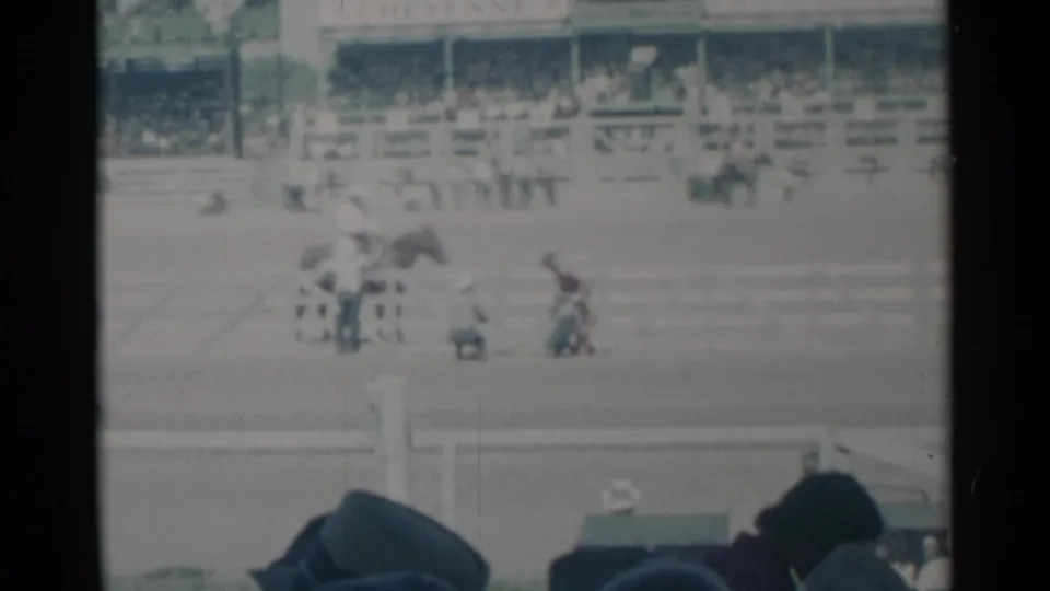 1960 Cheyenne rodeo arena view with horses and participants on the field, crowded grandstands behind, and audience heads at the bottom edge.