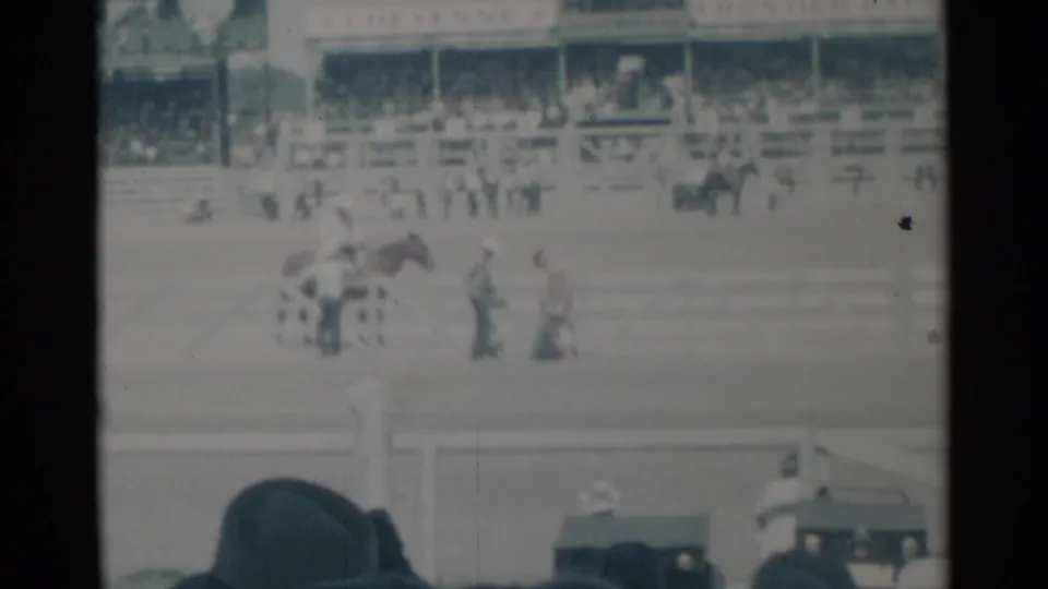 1960 Cheyenne rodeo scene showing a horse with nearby participants in the arena, viewed from behind spectators in the stands.