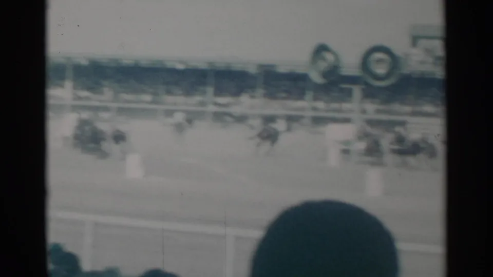 Soft 8mm view of multiple chariot teams racing in a Cheyenne rodeo arena, partly obscured by nearby spectators.