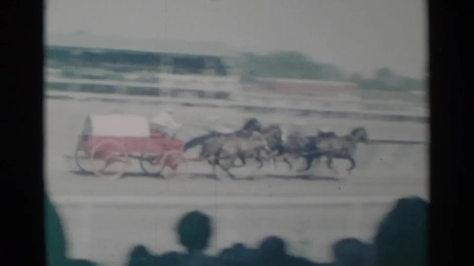 In faded 8mm footage from Cheyenne, several horses pull a red and white cart along the dirt track during a public race.