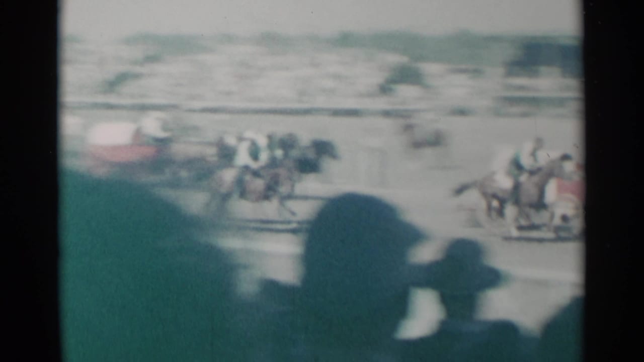 Jockeys and horses pass on the track with heads of spectators in foreground.