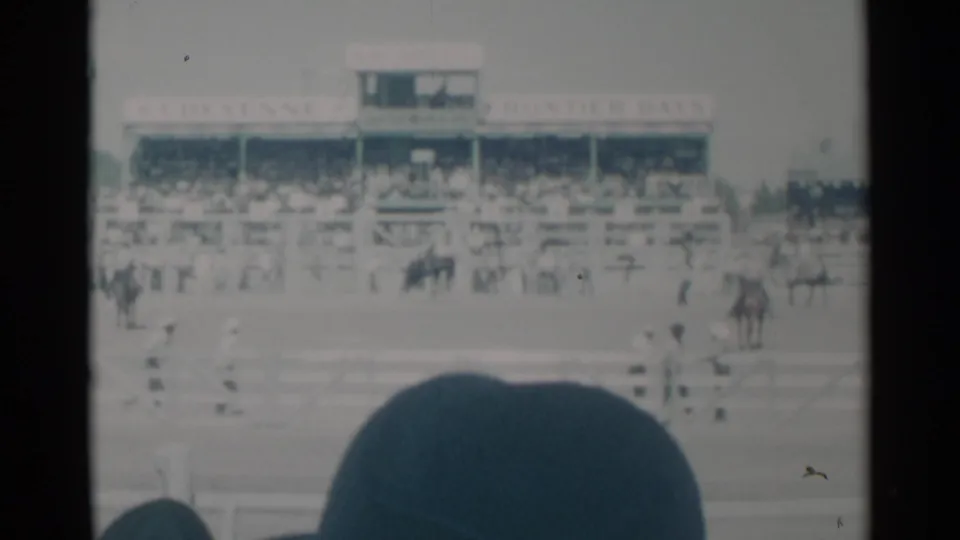 1960 Cheyenne rodeo view with a dark horse bucking in the arena, grandstands behind, and spectators partly blocking the frame.