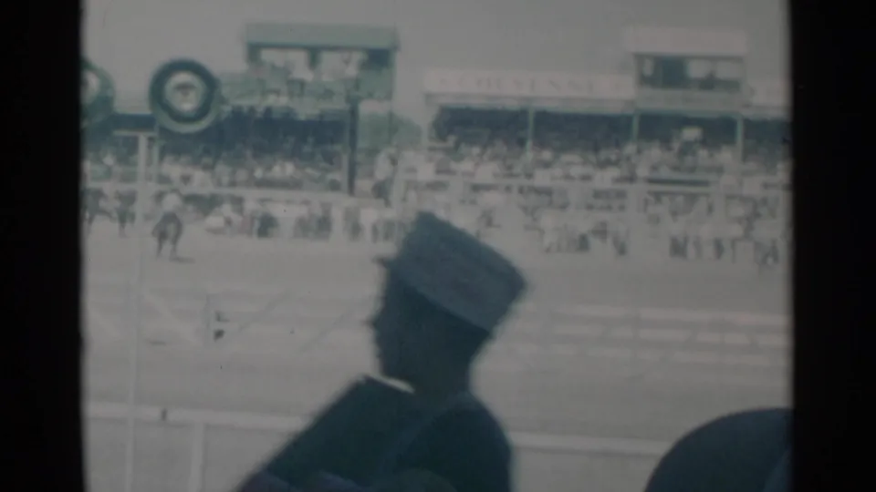 Audience view at a 1960 Cheyenne rodeo with a uniformed figure in silhouette, the grandstand, crowd, and arena horse activity behind.