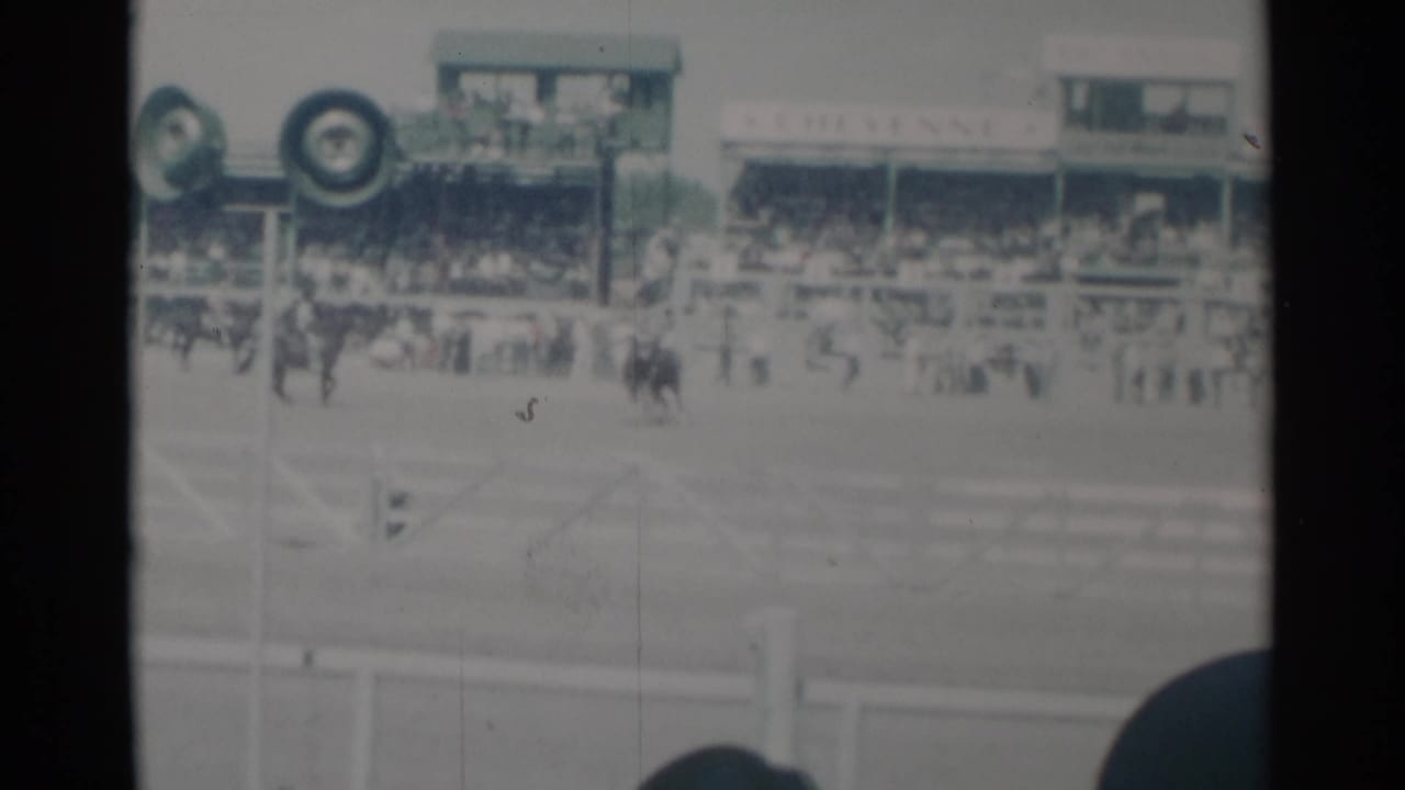 Horses and riders on a track in front of crowded grandstands, partly blocked by a spectator's head.