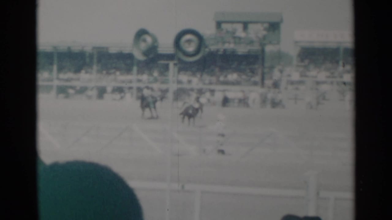 Riders on horseback move near the grandstand while a spectator's hat or head blocks the lower frame.