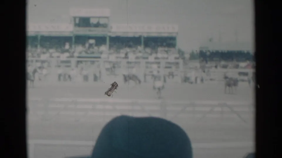 Soft 8mm frame from the 1960 Cheyenne event showing riders on the track, grandstand seating, railings, and a foreground spectator silhouette.