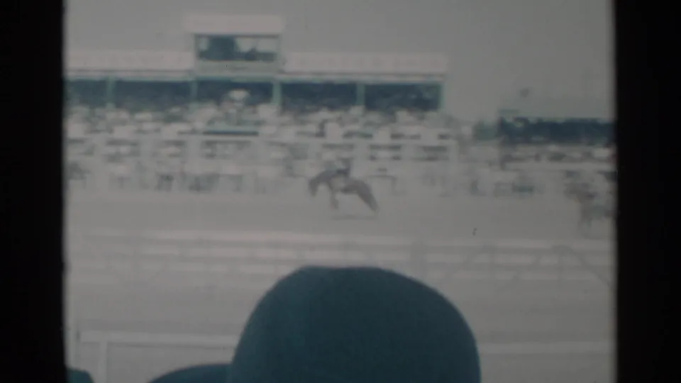1960 Cheyenne rodeo scene with a distant bronco rider crossing the dirt arena before crowded grandstands.