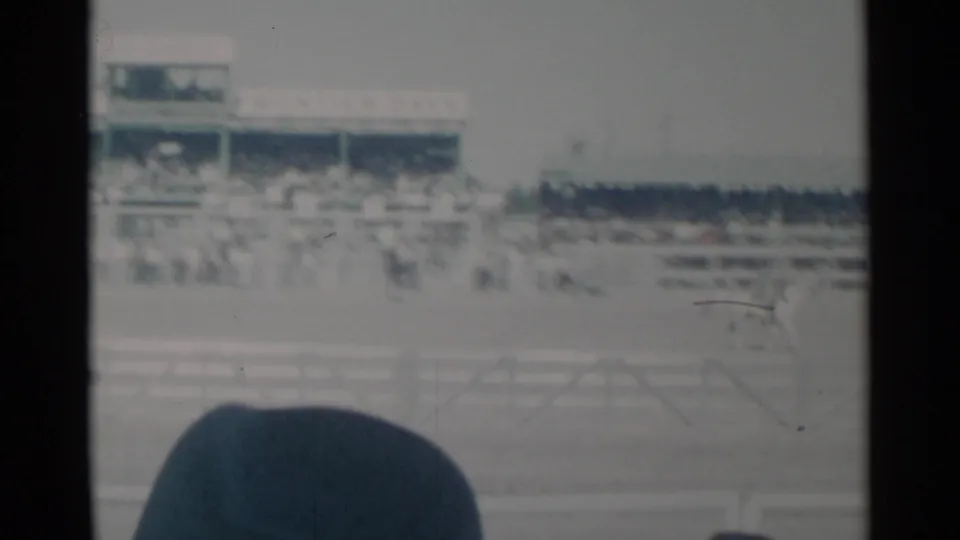 Soft 8mm view from the stands at a Cheyenne rodeo, with crowd, arena railings, and a large spectator hat in the foreground.