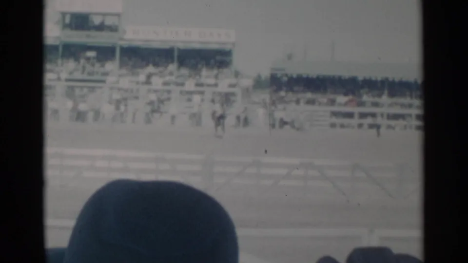 Cheyenne rodeo grandstands and arena railings seen from a spectator seat, with distant figures and a dark hat at the bottom of frame.