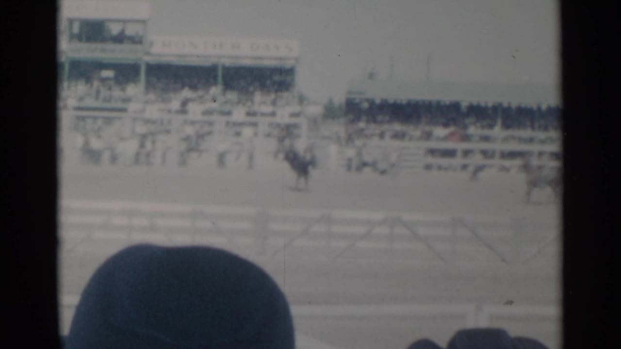 Mounted riders and horses cross a rodeo arena before crowded grandstands.