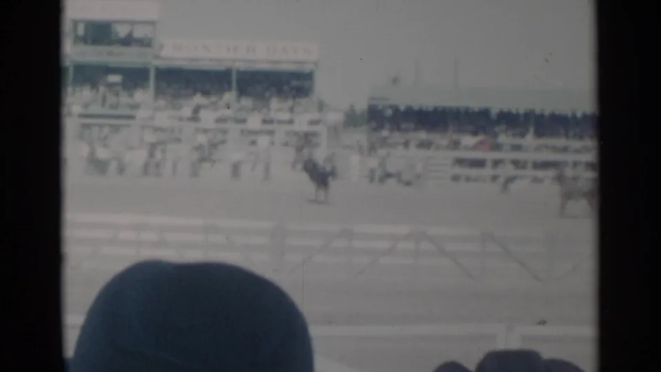 1960 Cheyenne, Wyoming rodeo arena view with riders on horseback, spectators in the stands, and a dark hat in the foreground.