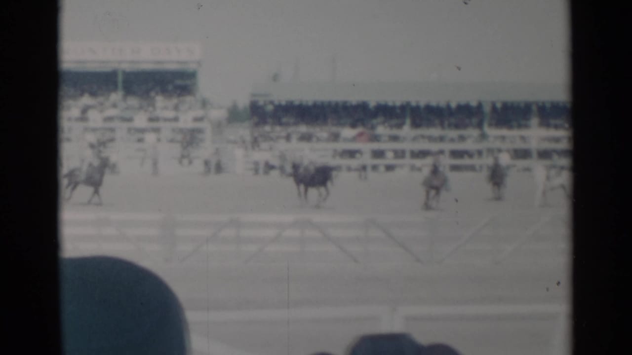 Riders on horseback pass along the arena with spectators behind them.