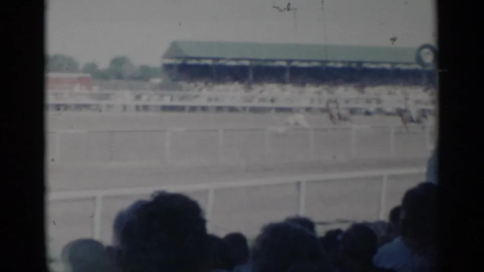 Cheyenne rodeo arena view from the stands, with foreground spectators, rail fencing, distant riders, and a long covered grandstand.