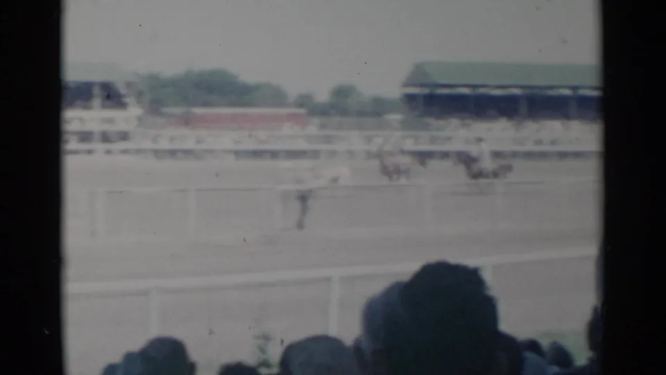 1960 Cheyenne rodeo arena viewed from the stands, with distant horseback riders and livestock beyond spectator silhouettes.