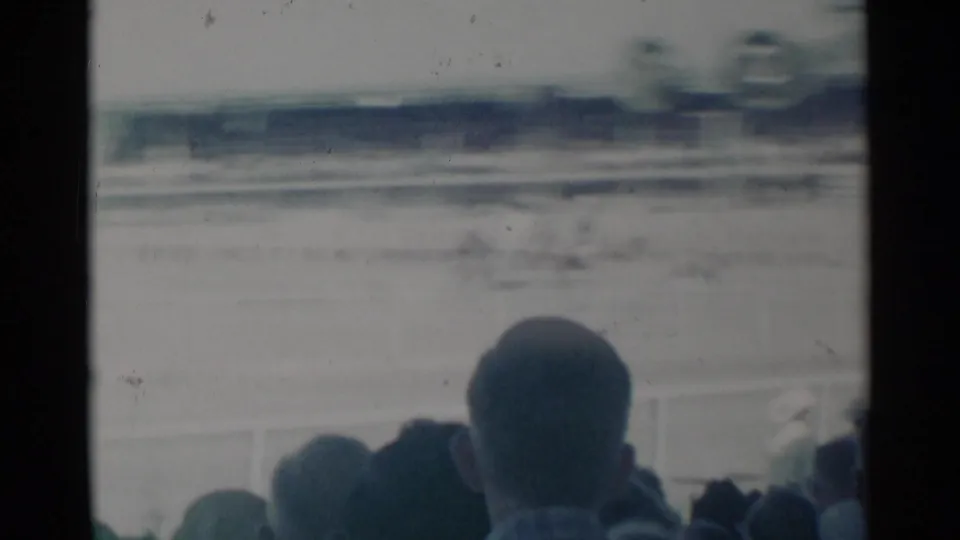From the spectator area, blurred 1960 Cheyenne rodeo action appears beyond railings and audience heads.