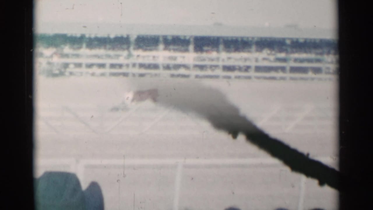 Dusty rodeo arena seen past dark film obstruction with white fencing and spectators in the background.