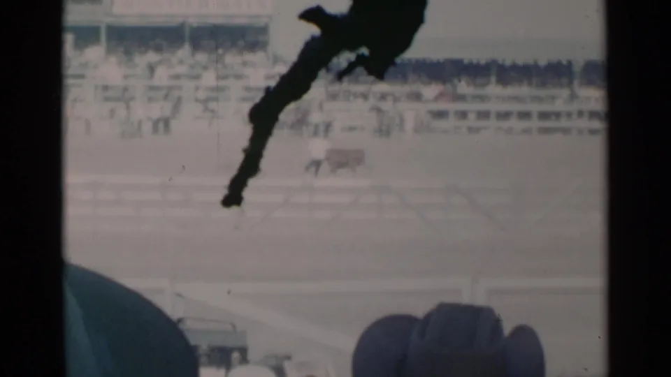 A 1960 Cheyenne rodeo scene shows distant livestock in the arena, white fencing, grandstand spectators, and a dark obstruction crossing the frame.