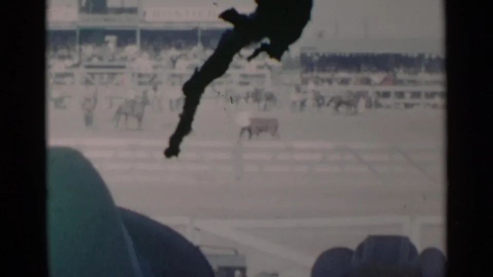 Cheyenne rodeo footage from 1960 shows a distant cow or steer and rider area in a dusty arena, with spectators behind white fencing.
