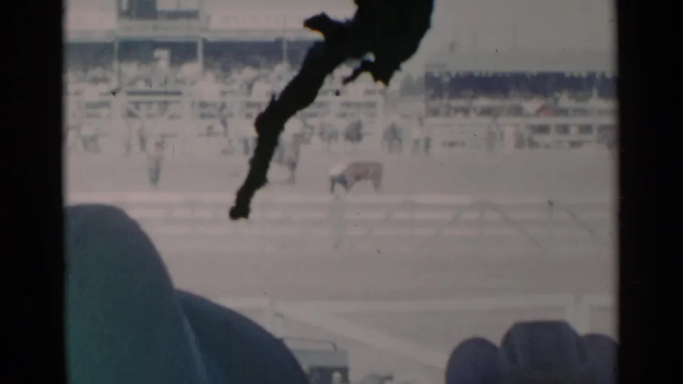 The closing frame holds on the 1960 Cheyenne rodeo arena, with distant livestock, grandstand spectators, white rails, and visible 8mm obstruction.