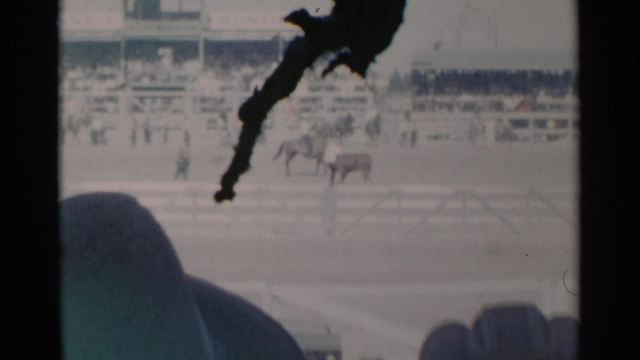 Rodeo arena viewed from stands with spectators in foreground and a dark obstruction across the frame.