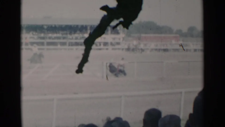 1960 Cheyenne rodeo scene with distant steer wrestling activity, arena railings, grandstand seating, foreground spectators, and soft 8mm film texture.
