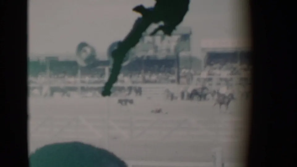 Wide 8mm view of the Cheyenne rodeo arena, with distant figures on the field, grandstand structures, foreground spectators, and heavy partial lens obstruction.