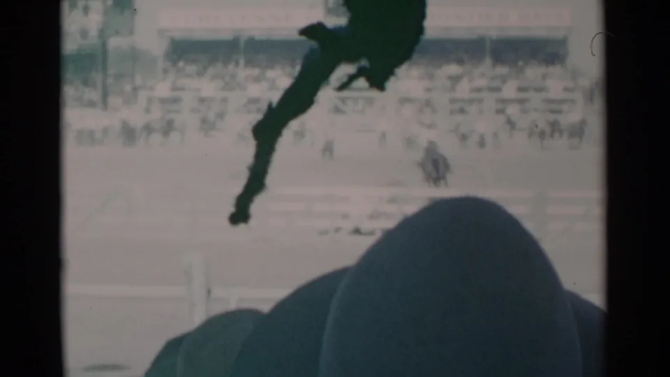 Cheyenne, Wyoming rodeo footage shows a foreground cowboy hat silhouette, arena fencing, and distant spectators or riders.