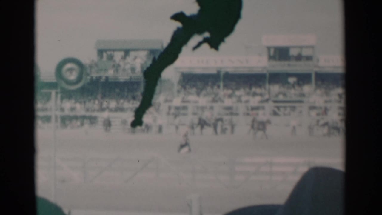 Cheyenne rodeo arena in 1960 with spectators in bleachers, riders in the ring, and a dark obstruction crossing the frame.