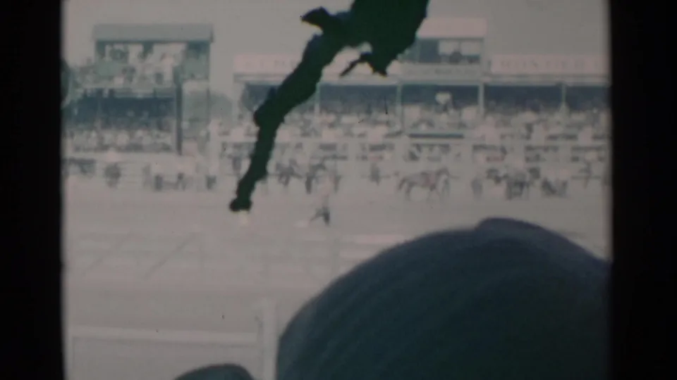 1960 home-movie view of a Cheyenne rodeo, with distant mounted riders, packed stands, and a spectator hat in front.