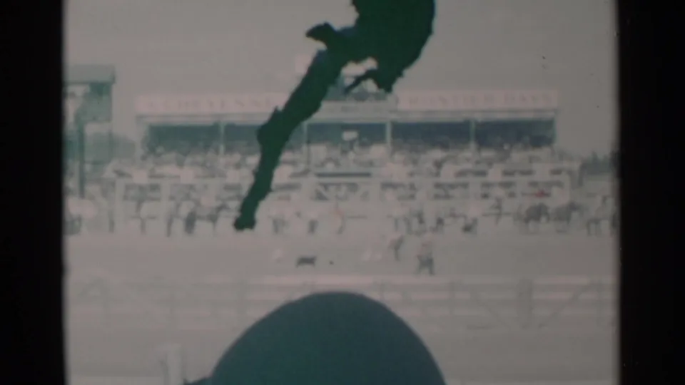 1960 Cheyenne rodeo footage showing a handler and small dog near ducks in the arena, with stands behind and a dark foreground obstruction.