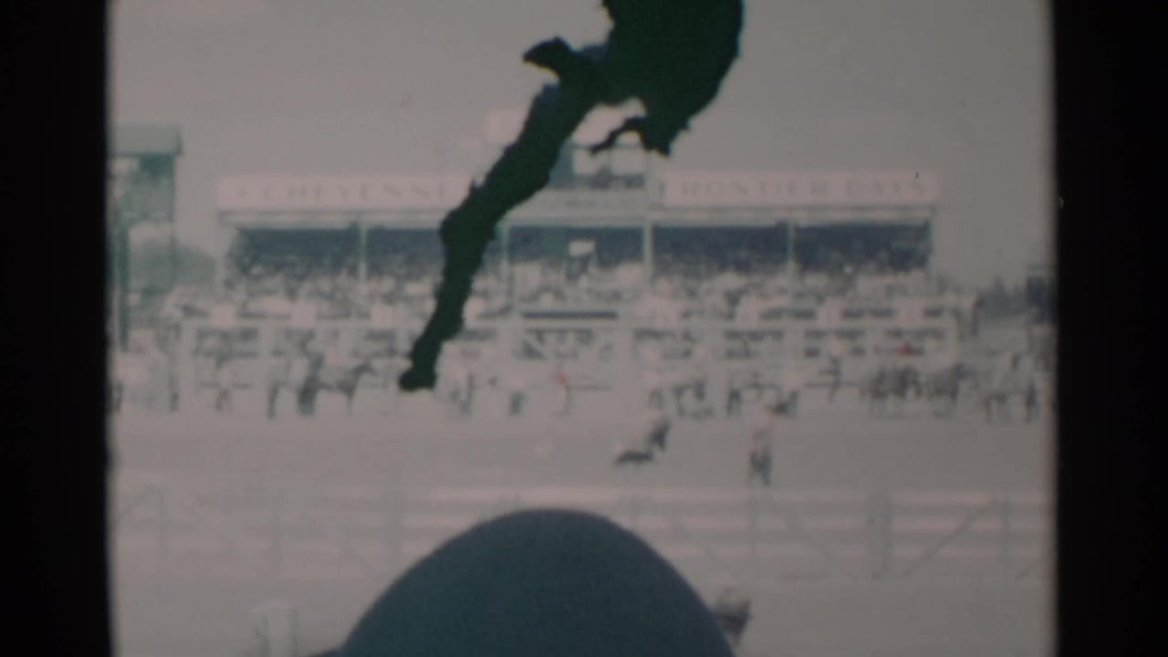 1960 Cheyenne rodeo grounds with spectators in the stands, arena figures, and a dark foreground shape partly blocking the 8mm frame.