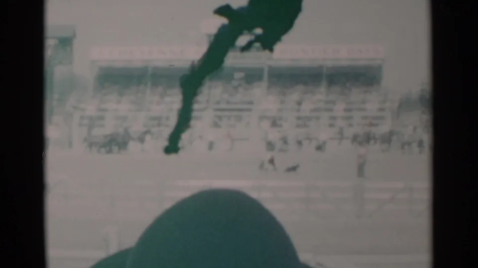 Faded 8mm frame of the Cheyenne rodeo grounds, with a packed grandstand, scattered arena figures, fencing, and a dark foreground obstruction.