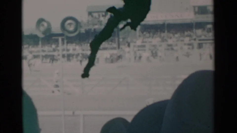 Cheyenne rodeo grounds with spectators, circular arena markers or equipment, distant figures, and dark foreground shapes along the 8mm frame edge.