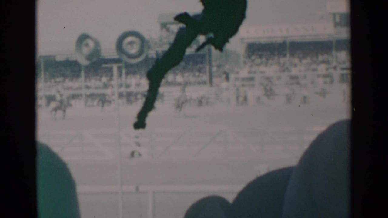 Bucking horse and rider in a rodeo arena with grandstands and foreground spectators.